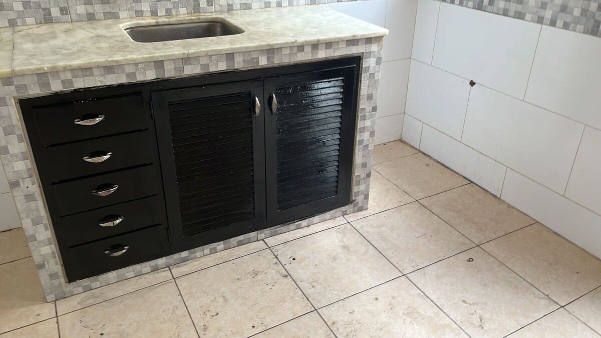 A kitchen area is presented with a light-colored marble countertop and a stainless steel sink. Below, a black cabinet with horizontal slats features several drawers, providing storage space. The tiled floor is light beige, and the walls are adorned with white tiles accented by a gray mosaic border.