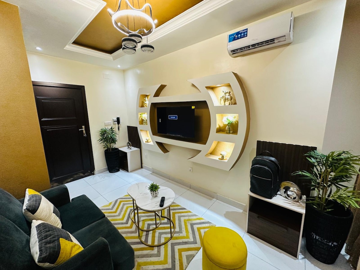A modern living area features a stylish TV unit with ambient lighting, showcasing decorative items. A cozy sofa with patterned cushions complements a round coffee table and a vibrant yellow accent pouf. A small indoor plant adds a touch of greenery, while air conditioning is visible for comfort.