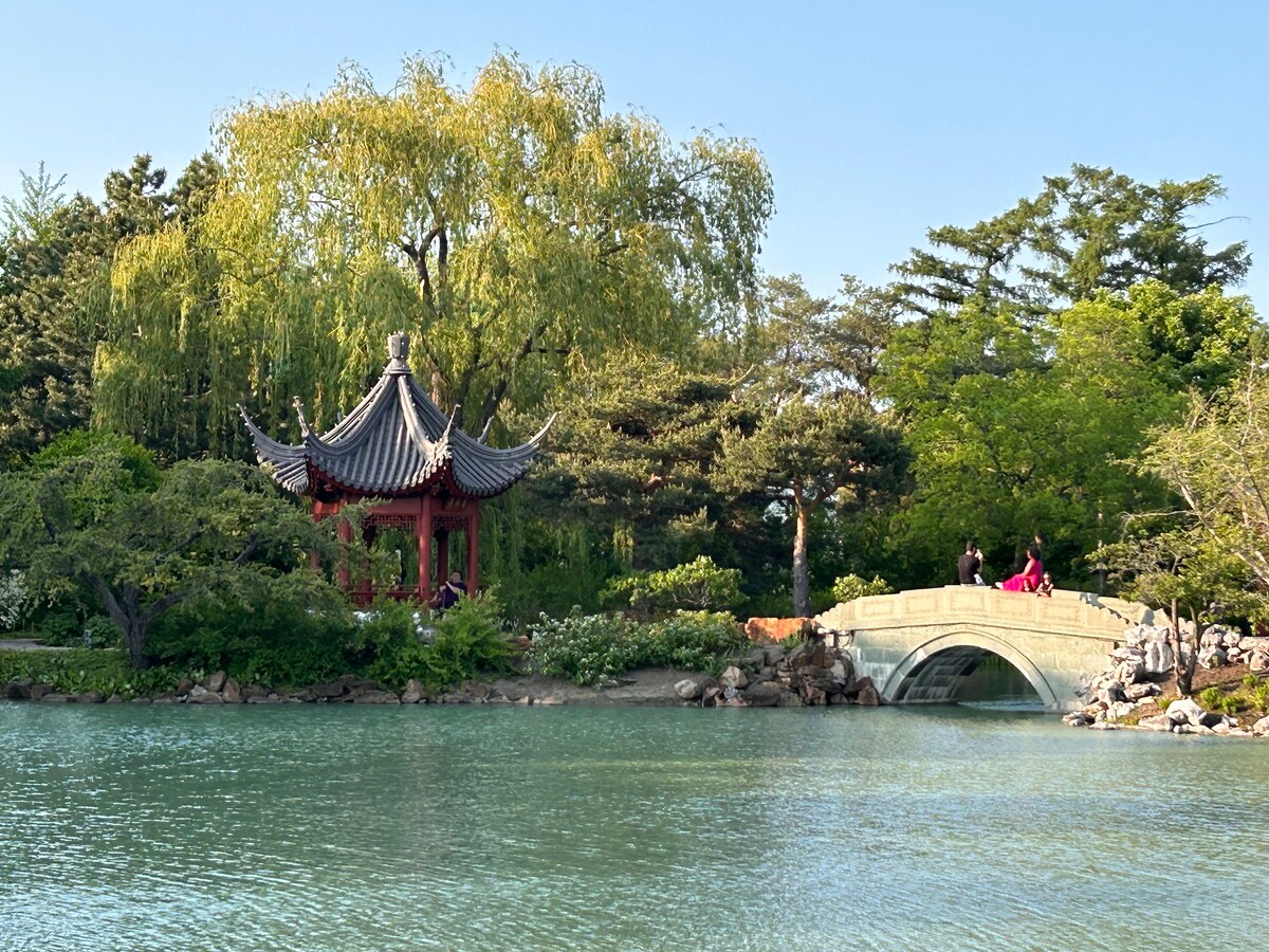 A serene landscape captures a picturesque garden scene. A traditional red pavilion is bordered by lush green trees, with a stone bridge arching over a still turquoise body of water reflecting the clear sky.