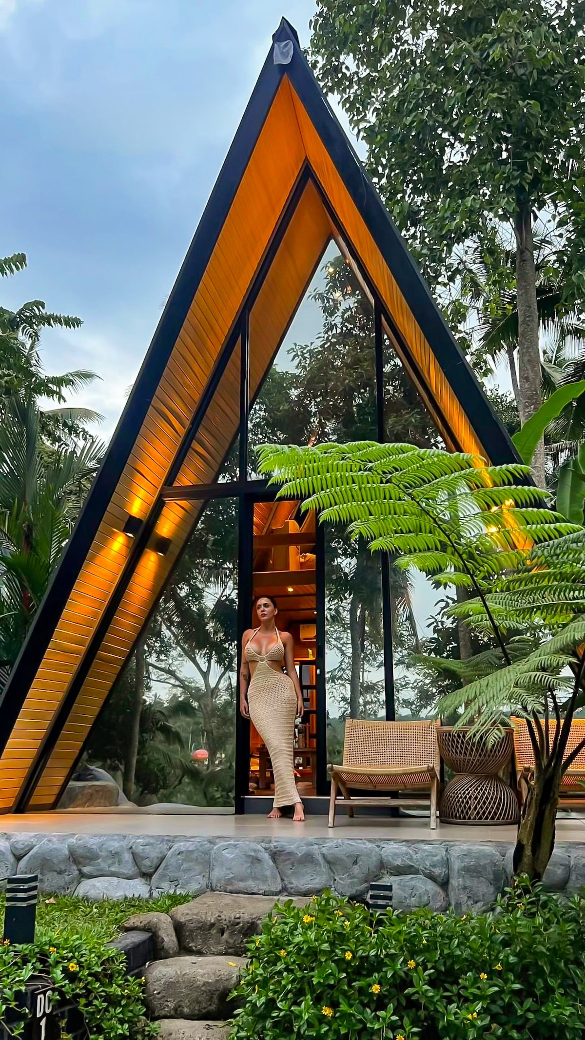 An A-frame villa with large glass panels rises amidst lush greenery, showcasing warm lighting emanating from inside. The entrance is framed by tropical plants, and a cozy seating area with wicker furniture is visible on the patio.