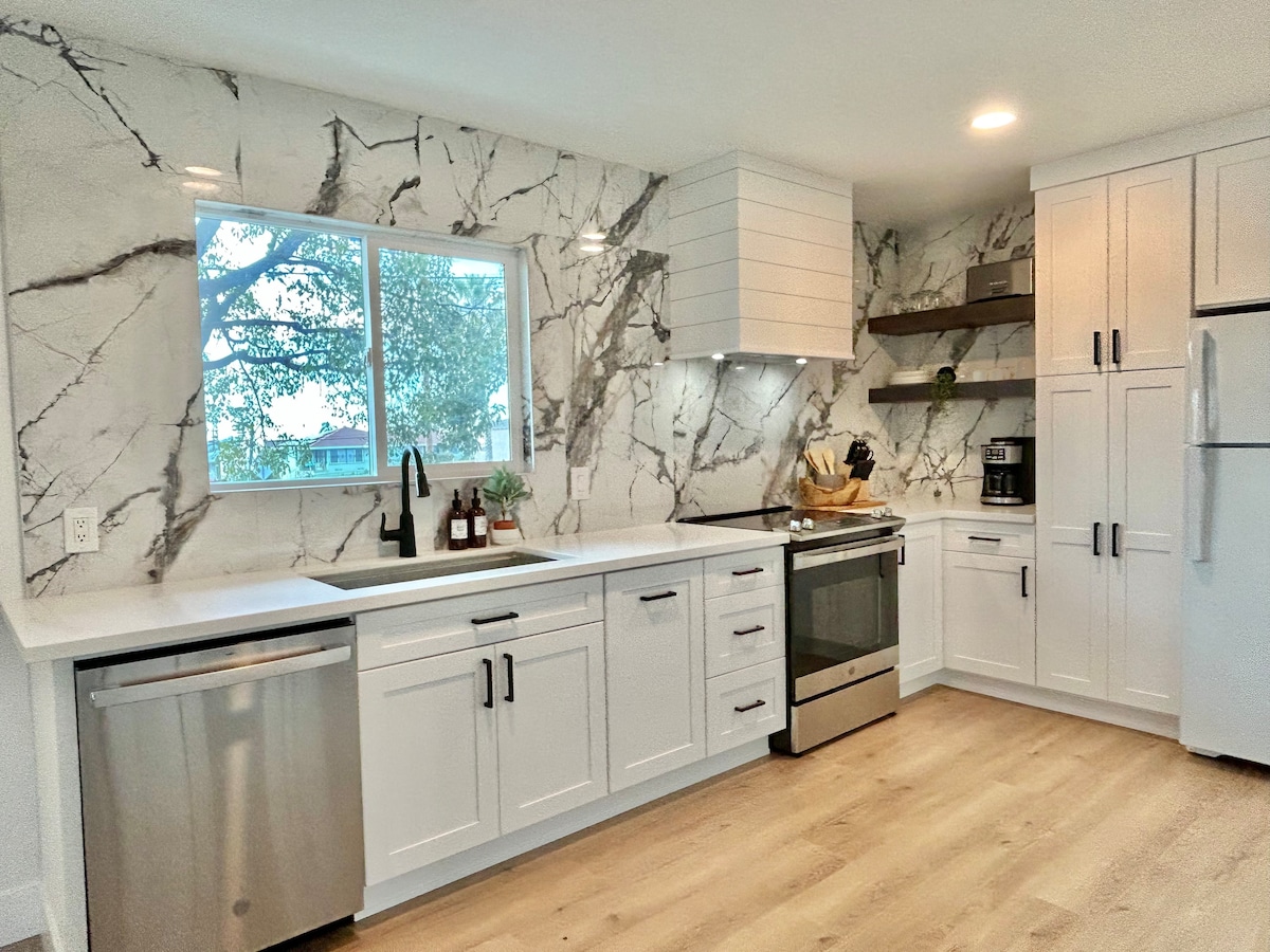 A modern kitchen features sleek white cabinetry with black hardware and marble-patterned walls. A stainless steel refrigerator and dishwasher are positioned for convenience. Natural light fills the space through a window, providing a view of greenery outside. Open shelving is displayed with decorative items.