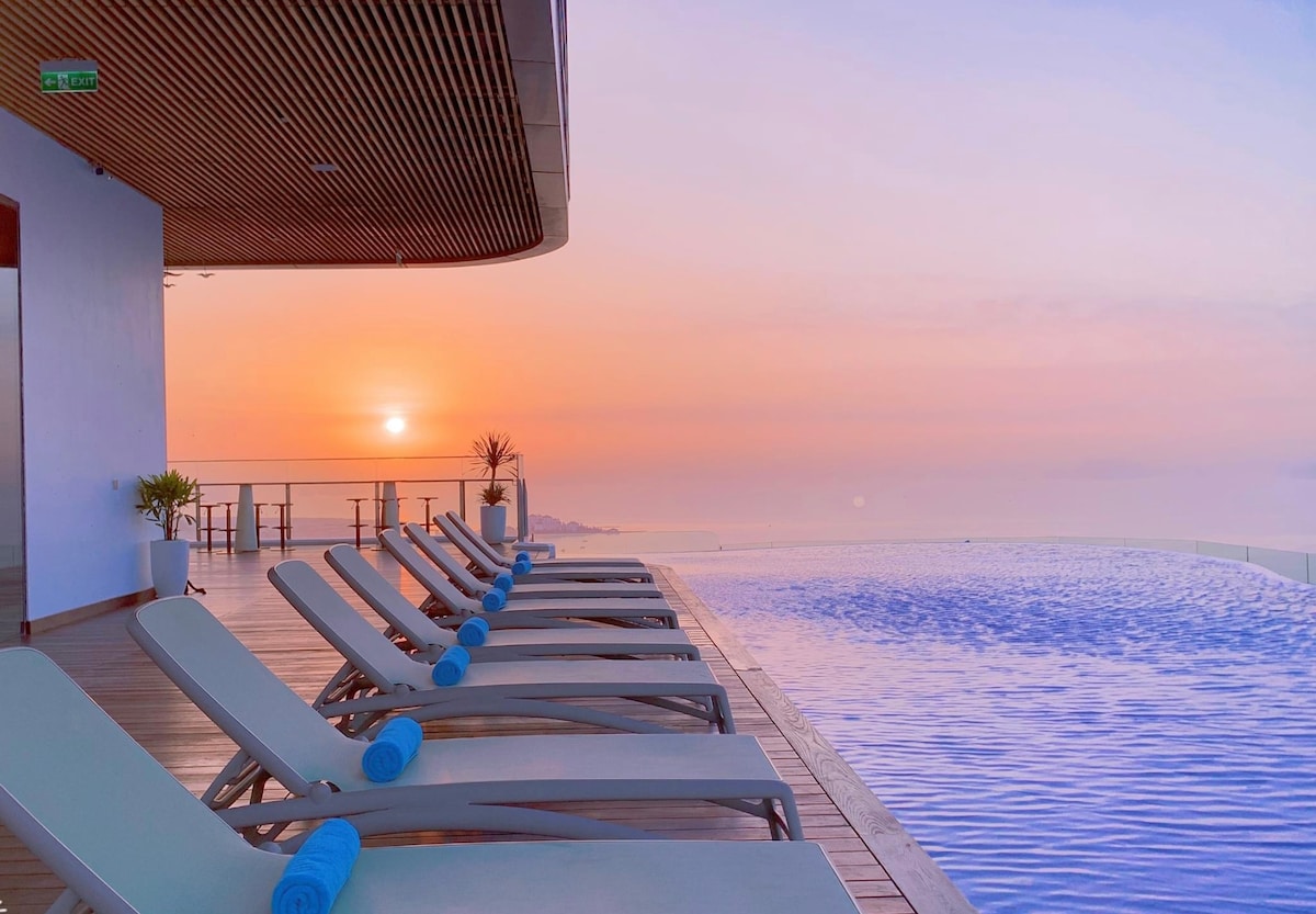 An infinity pool is framed by lounge chairs arranged neatly along the deck. A serene view of the sunrise reflects off the water, creating a peaceful ambiance. The warm colors of the sky enhance the tranquil setting, inviting relaxation.