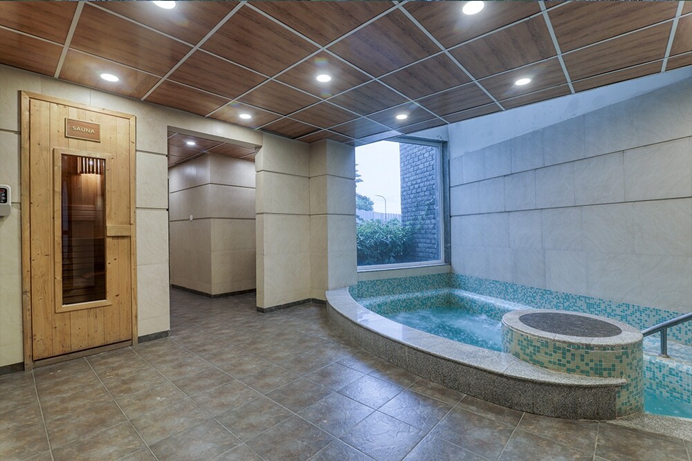 An indoor space features a small circular pool with mosaic tiling, illuminated by recessed lighting. A wooden sauna room is visible on the left, with large windows allowing natural light to fill the area. Neutral-toned walls and floor tiles create a calming environment.
