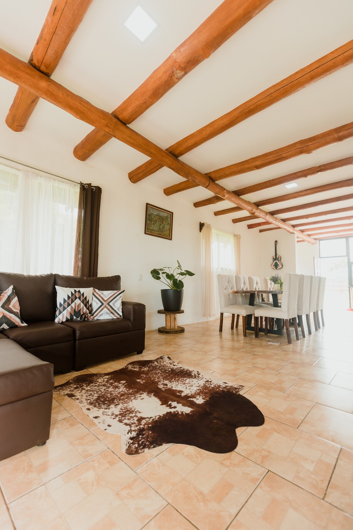A cozy living area features a brown leather sectional sofa adorned with patterned cushions. A light-colored dining table with white chairs is positioned nearby. The space is illuminated by natural light through large windows, and a patterned area rug adds texture to the tiled floor.