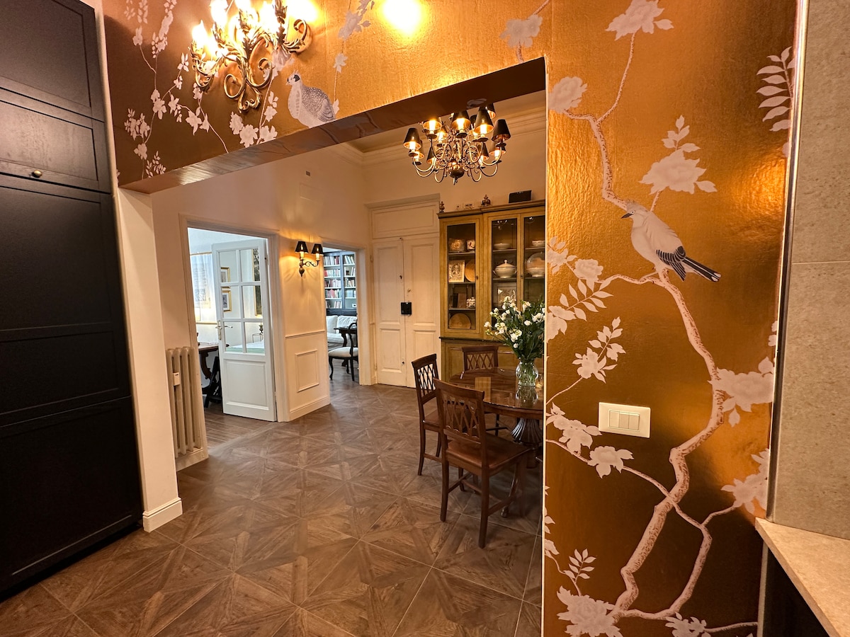 A dining area features a wooden table with four chairs, set against a wall adorned with gold and floral wallpaper. A chandelier hangs from the ceiling, providing soft illumination. Light enters through an arched doorway leading to an adjoining room with additional seating.
