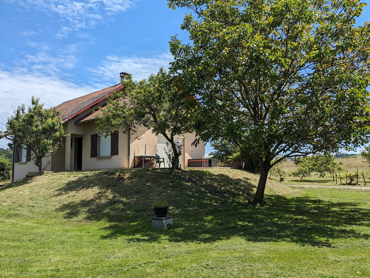 A charming house is set on a gentle hill, surrounded by lush green grass and trees. The structure features multiple windows and a covered patio furnished with a table and chairs, creating an inviting outdoor space for relaxation.