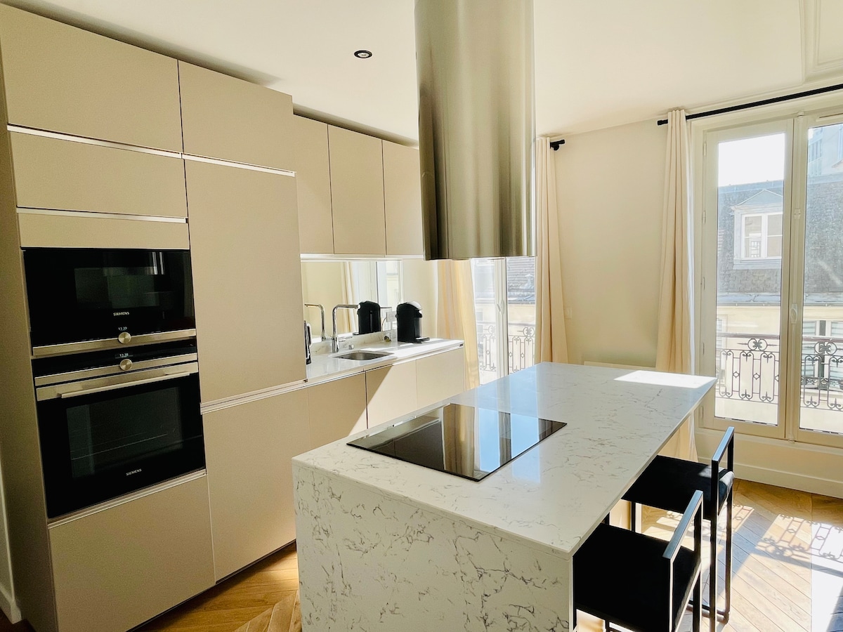A modern kitchen features sleek cabinetry with integrated appliances and a spacious island. A cooktop is situated on the island, accompanied by two black chairs. Natural light streams in through large windows adorned with light curtains, illuminating the minimalist design.