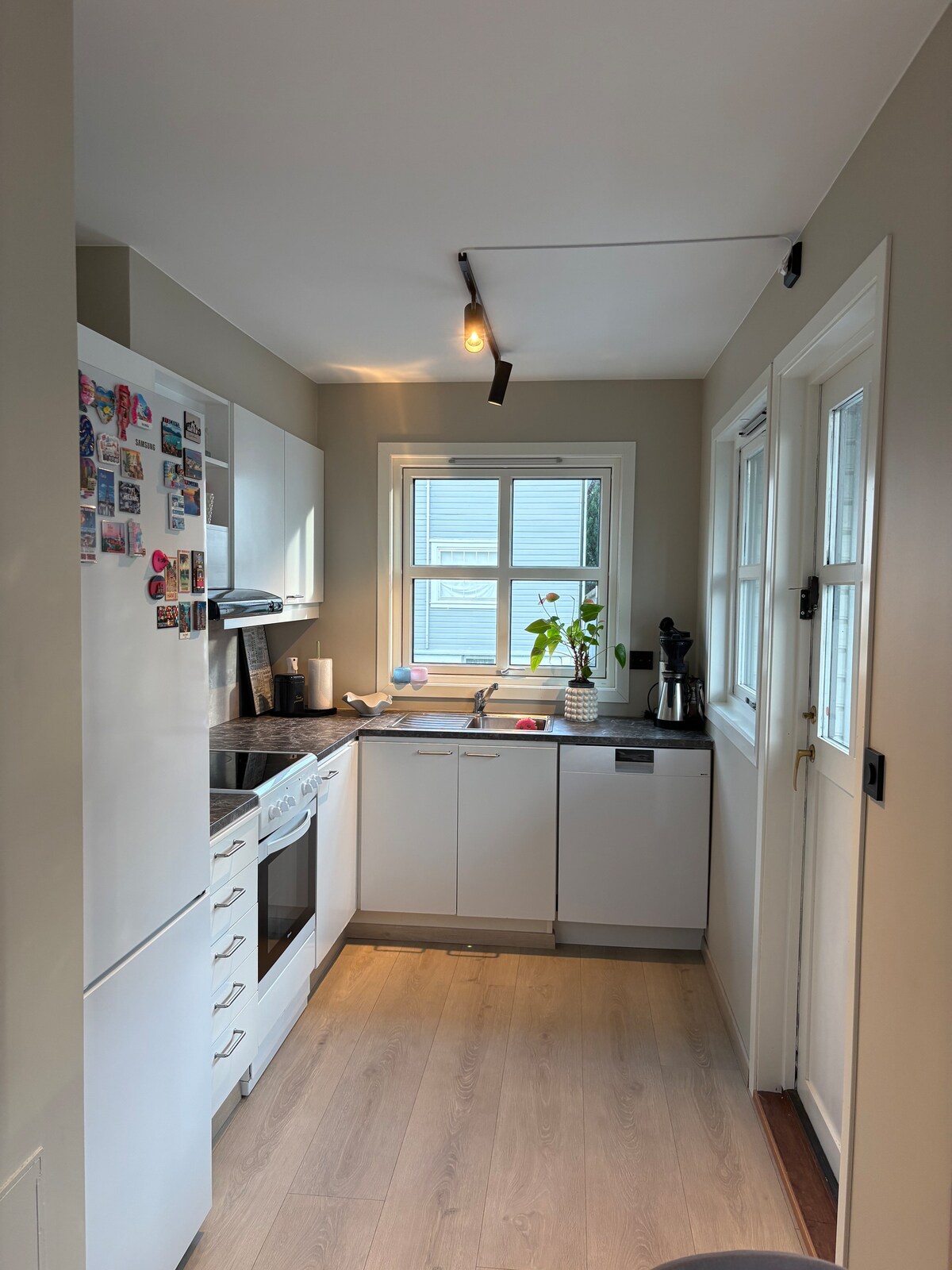 The kitchen features modern appliances including a refrigerator, oven, and dishwasher. A window allows natural light to brighten the space, complemented by minimalistic cabinetry. A small table is placed nearby, and decorative touches, such as a plant, enhance the environment.