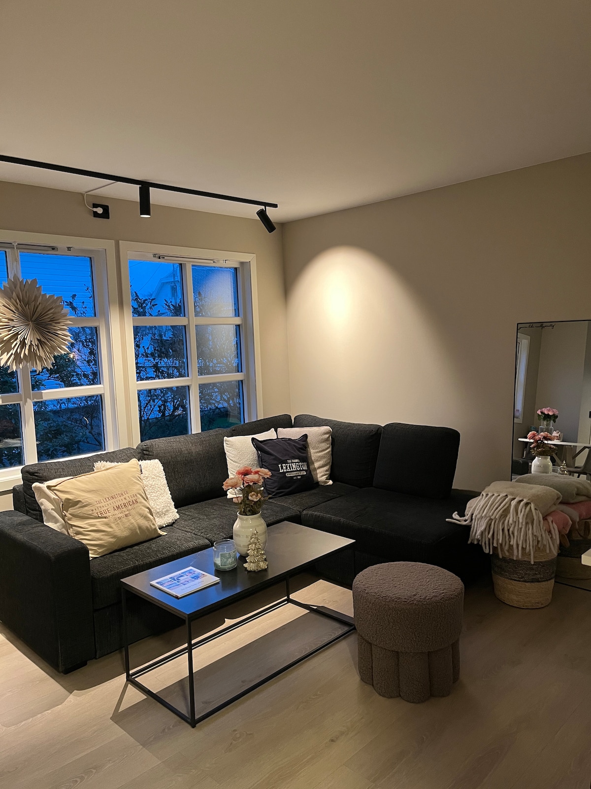 A modern living area features a dark sectional couch adorned with various cushions. A small round ottoman sits nearby, while a minimalist coffee table holds decor items. Natural light filters through multiple windows, highlighting a neutral color palette and creating a calm ambiance.