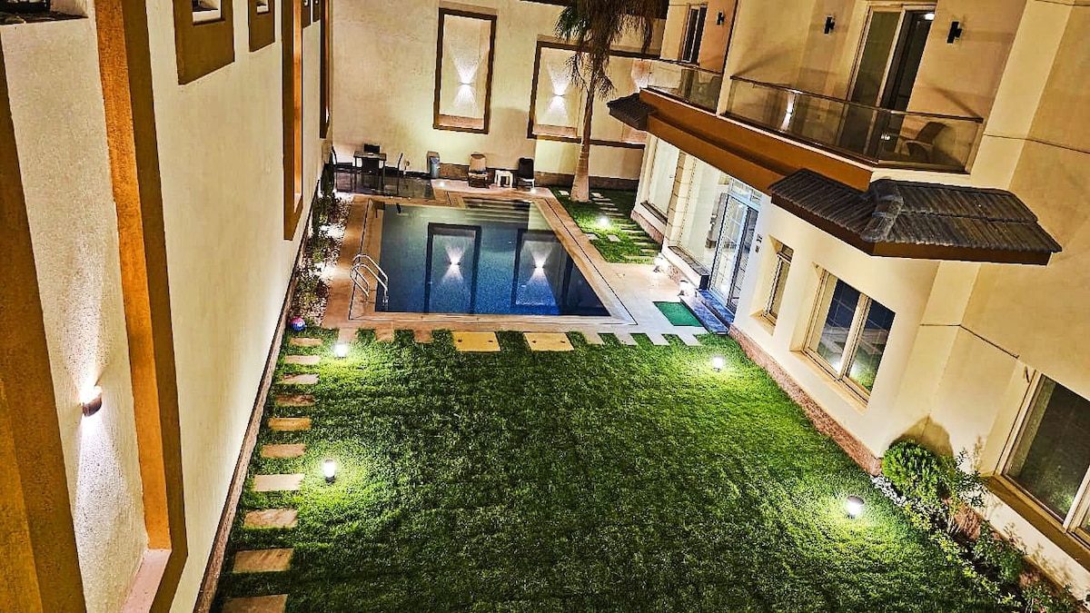 A well-maintained outdoor space features a rectangular pool surrounded by manicured lawn and stepping stones. Lounge chairs are positioned near the pool, and soft lighting enhances the area, providing a serene ambiance in the evening.