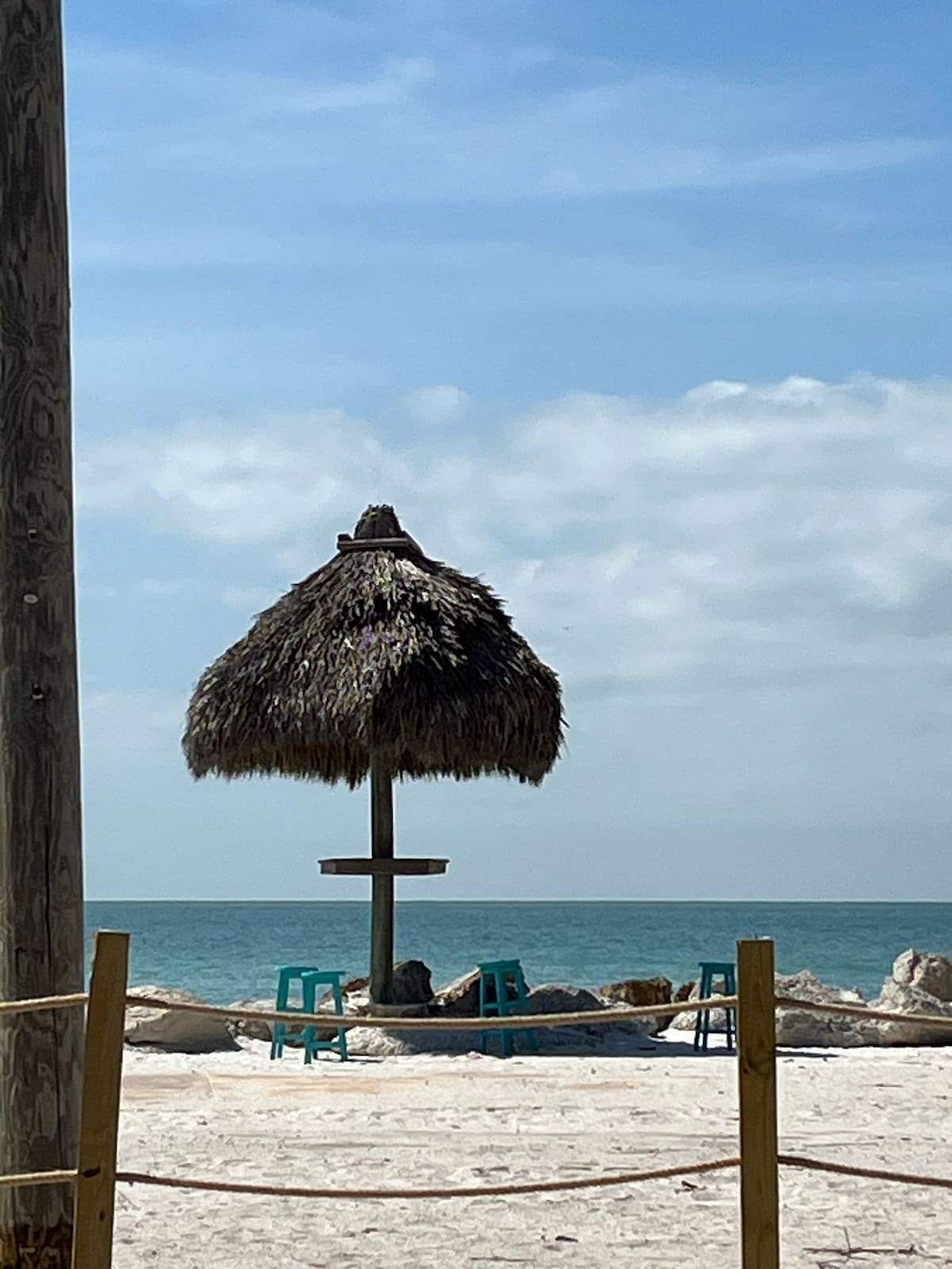 A thatched gazebo stands on the beach, providing shade near the water's edge. Two green chairs are positioned in front of the gazebo, facing the calm blue ocean. The sandy beach is visible, along with a clear sky dotted with soft clouds.