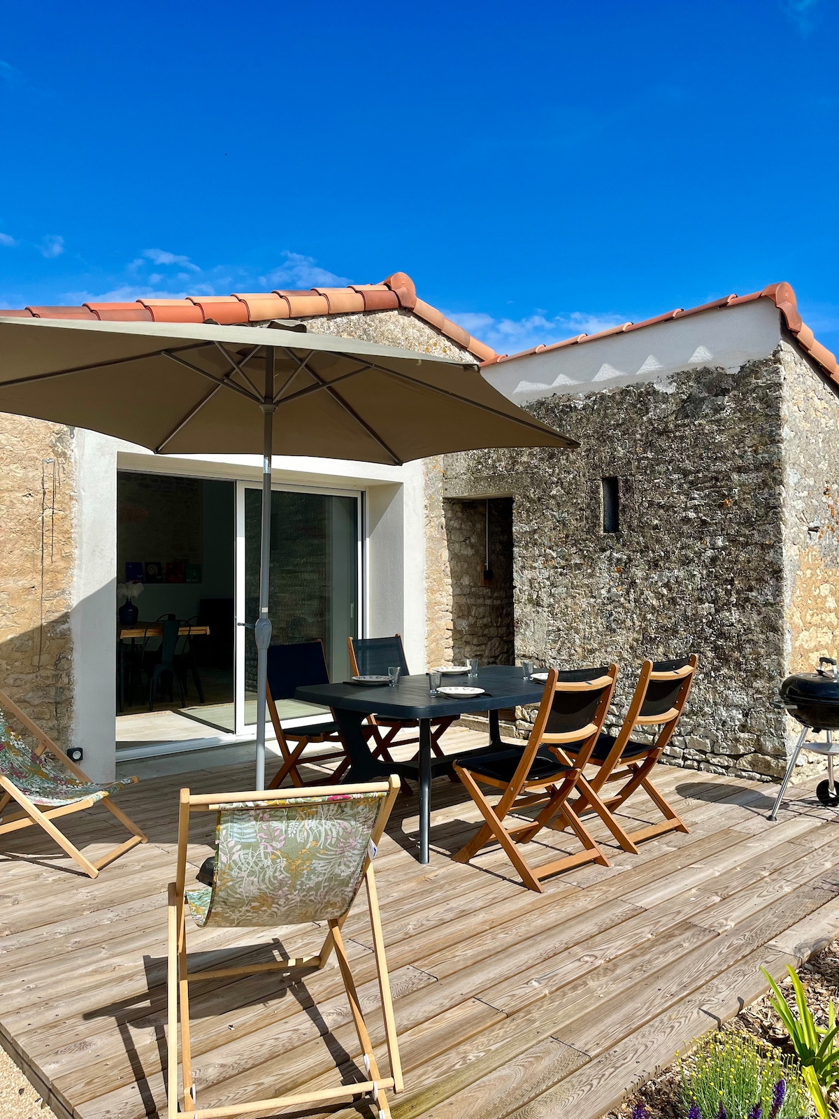 An outdoor terrace features a wooden deck with a dining table surrounded by chairs. A large umbrella provides shade, while a barbecue stands near the edge. The backdrop includes a stone wall and large glass doors leading into the living space, under a clear blue sky.