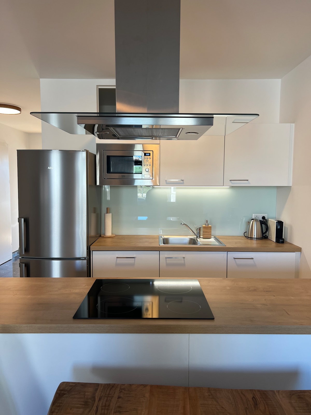 The kitchen features a sleek layout with modern appliances, including a refrigerator, microwave, and kettle. A stainless steel hood hangs above a glass-ceramic cooktop, while a wooden countertop provides ample workspace, complemented by a neutral backsplash and soft lighting.