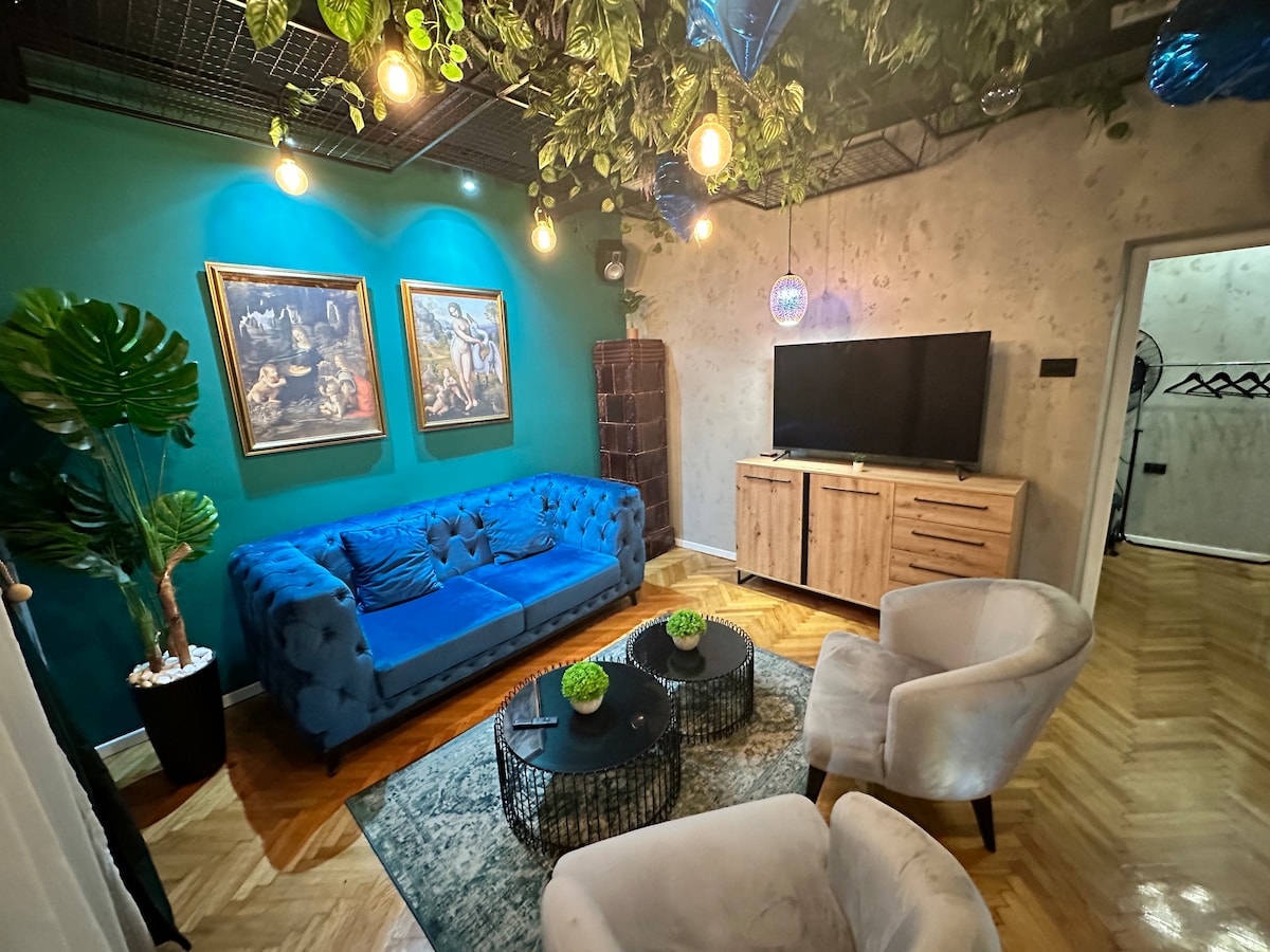 A living area features a blue velvet sectional sofa paired with two cream-colored armchairs. A wooden console holds a flat-screen television, while two round coffee tables sit atop a patterned rug. Wall art and greenery add a touch of elegance to the environment.