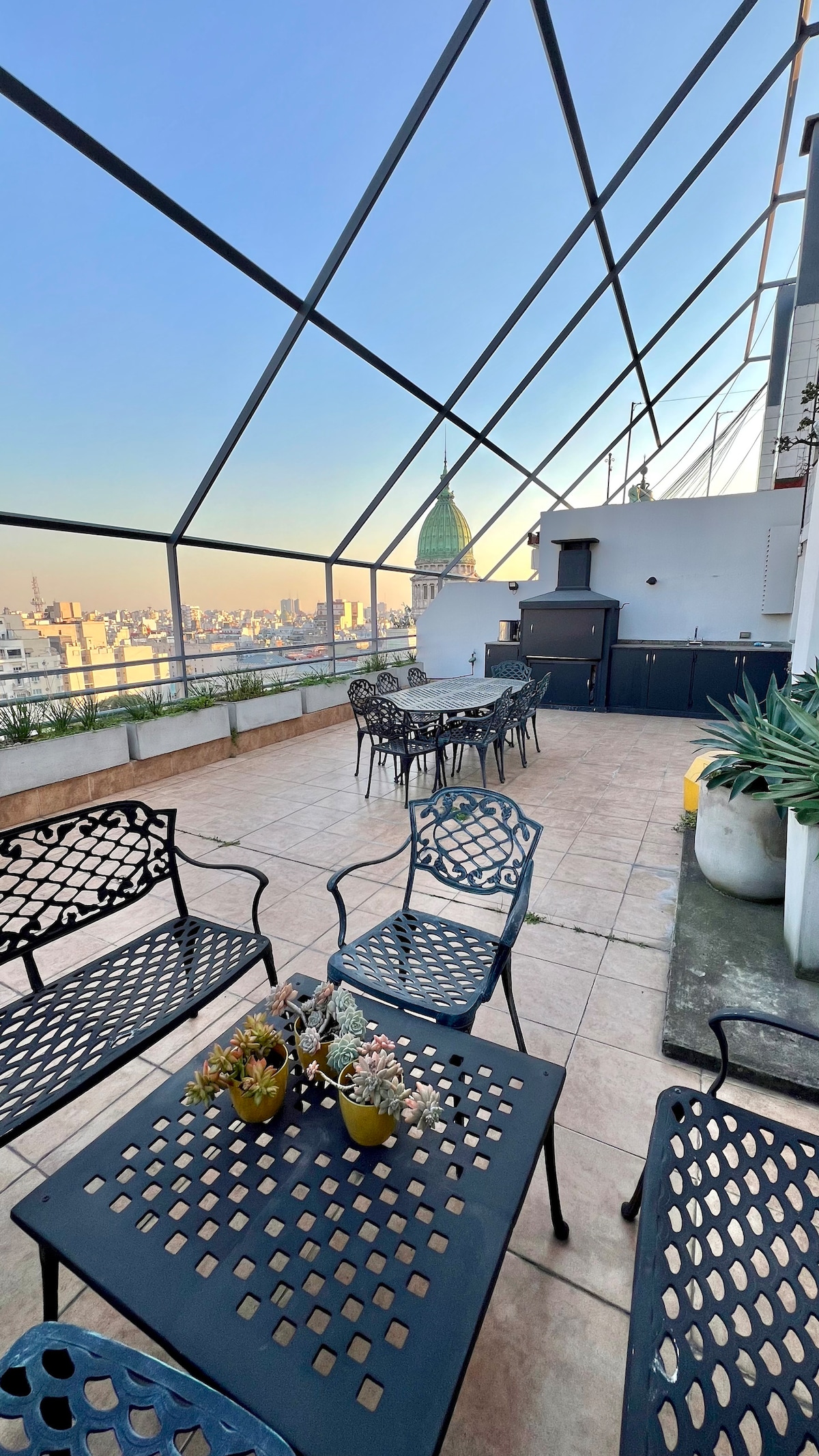 The spacious rooftop terrace features multiple black metal tables and chairs arranged for gatherings. A barbecue area is present, and potted plants add greenery. The stunning view includes the iconic dome of the Congress building, framed by a clear sky.