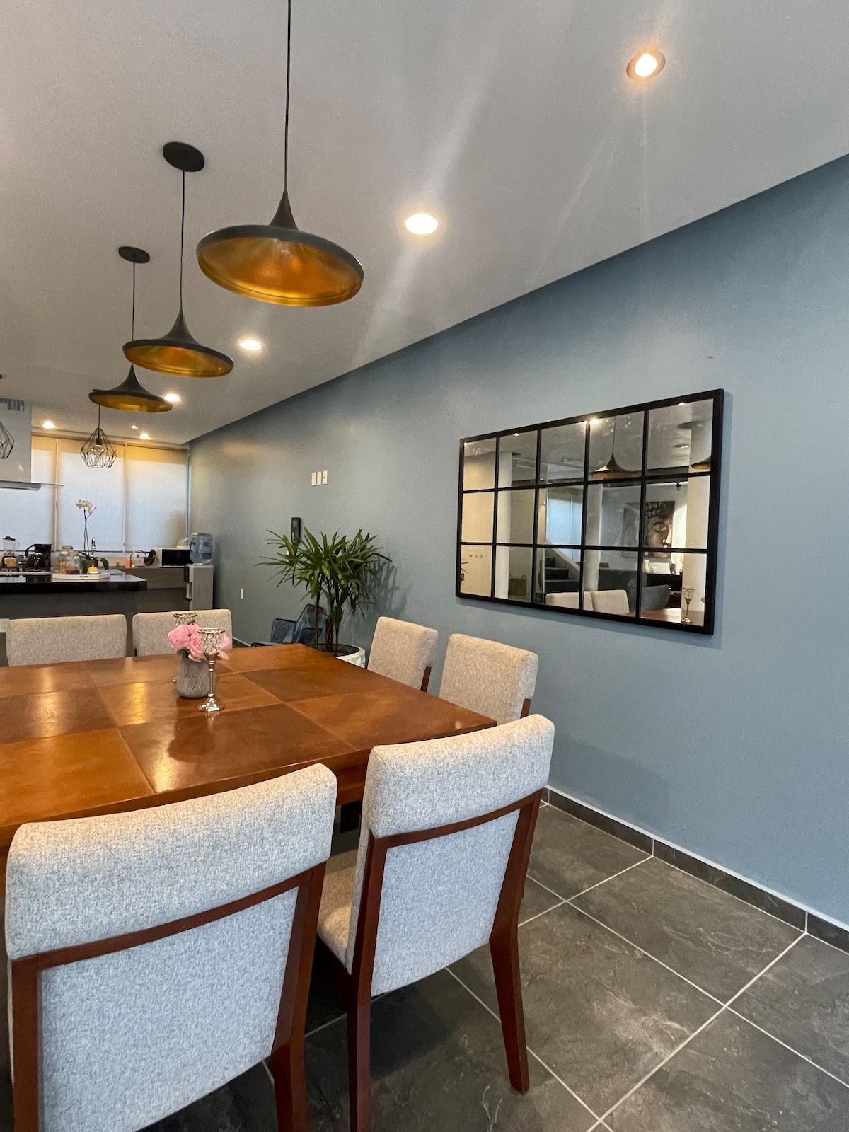 A dining area is presented with a wooden table surrounded by upholstered chairs. Three pendant lights hang above, illuminating the space. A decorative mirror mounted on the wall reflects the room's ambiance, enhancing the sense of openness.