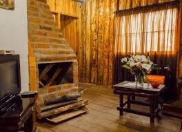 A living area features a rustic brick fireplace accented by wooden paneling. A small wooden table is paired with a chair, and a bouquet of flowers adds a natural touch. Natural light filters through the window, partially covered by sheer curtains.