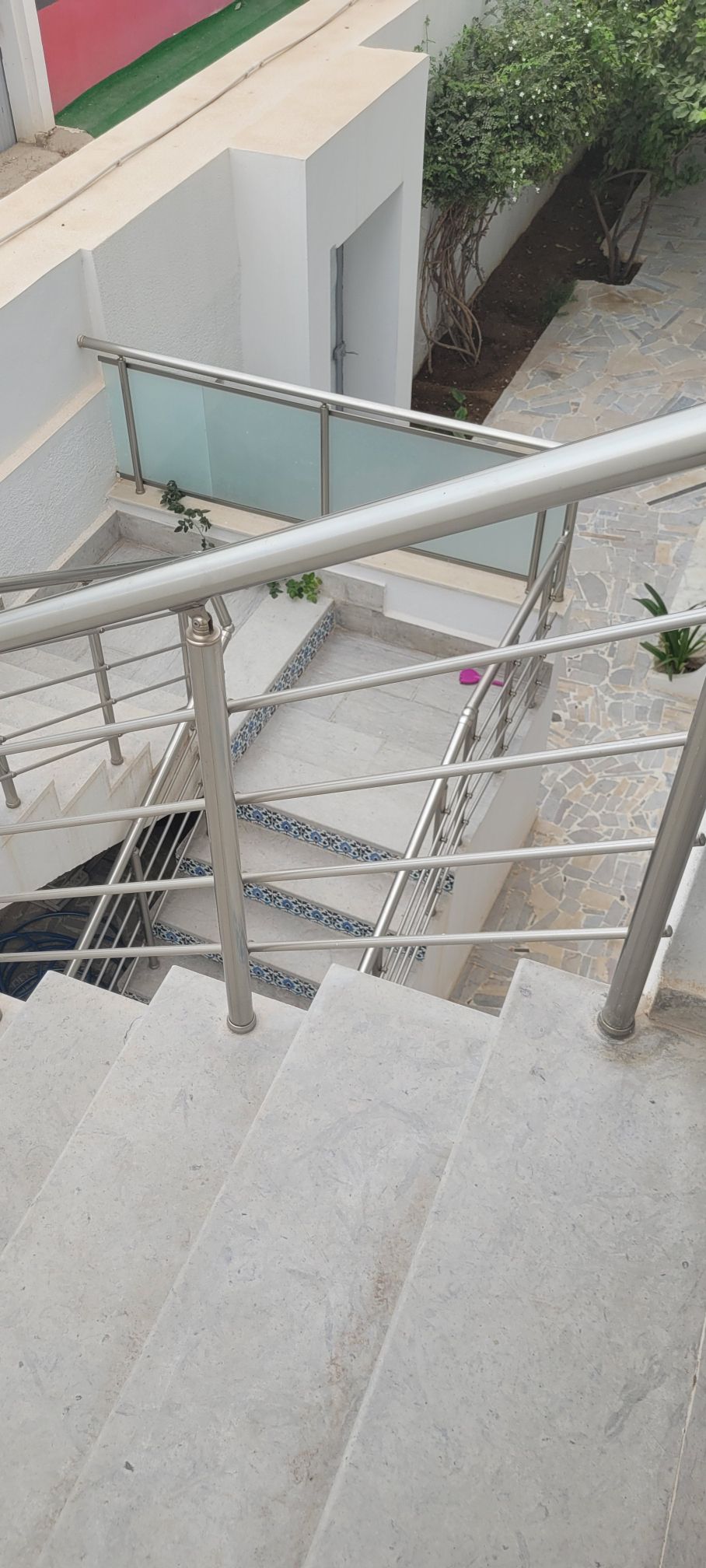 A well-maintained staircase with a stainless steel railing leads down to a stone pathway. The steps are bordered by a glass partition, and green plants are visible along the sides. Natural light illuminates the area, highlighting the clean design and materials.