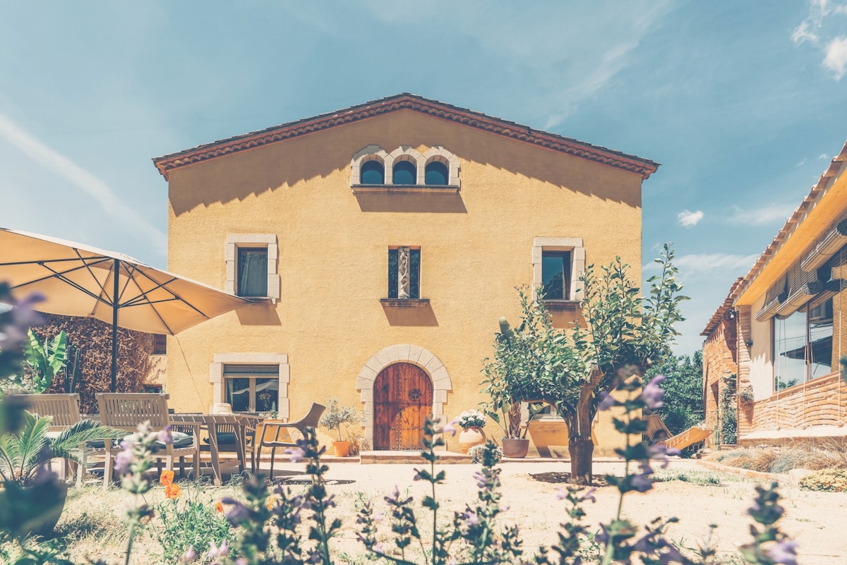 A rustic yellow farmhouse with a tiled roof is framed by blossoming plants in the foreground. The entrance features a decorative wooden door, flanked by two windows. A shaded outdoor seating area with a large umbrella and dining set is visible to the left.