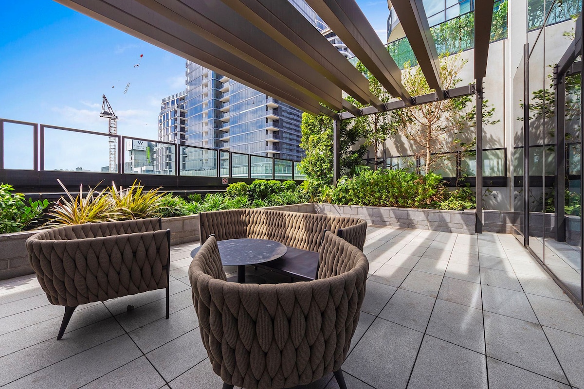 A spacious outdoor balcony is furnished with two stylish wicker chairs arranged around a small table. Lush greenery and low-maintenance plants enhance the area, which is framed by modern buildings and a clear blue sky. Natural light streams across the tiled floor.