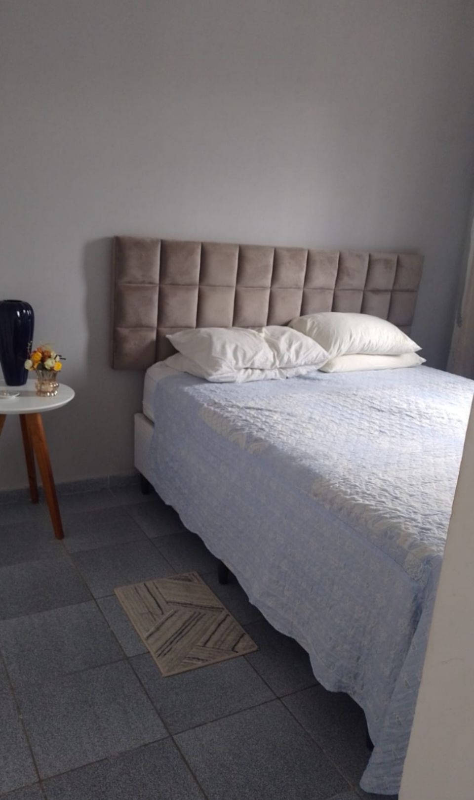 A sleeping area features a spacious bed with a light blue quilted coverlet and a tufted headboard. Two white pillows are placed at the head, and a small round table with a vase and flowers is positioned nearby.