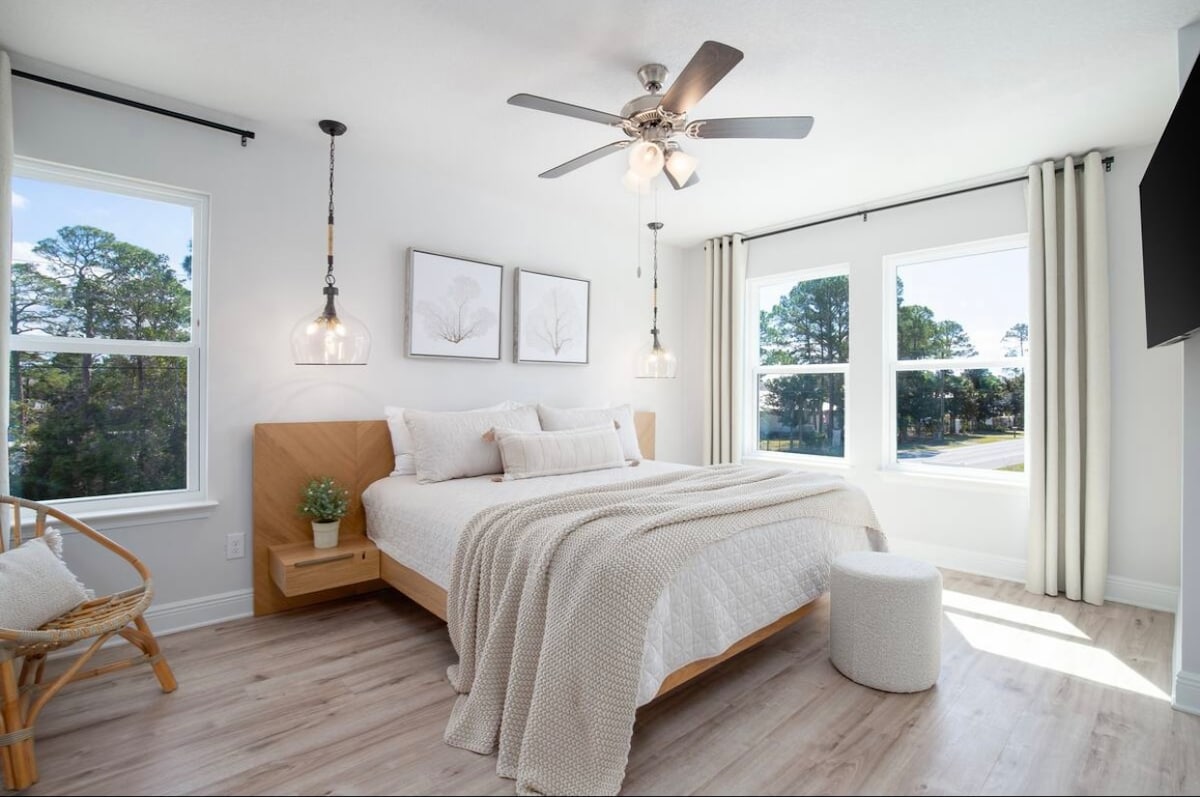 A bright bedroom features large windows that allow natural light to illuminate the spacious bed. Light-colored walls and soft bedding create a serene environment. A ceiling fan and unique light fixtures enhance the space, while a small stool and a rattan chair provide additional seating.