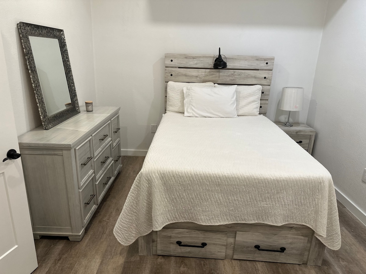 A compact sleeping area features a full-size mattress, framed by a rustic headboard. A dresser and nightstand in light wood finish provide ample storage. Natural light enters through a window, and a decorative mirror adds a touch of style to the room.