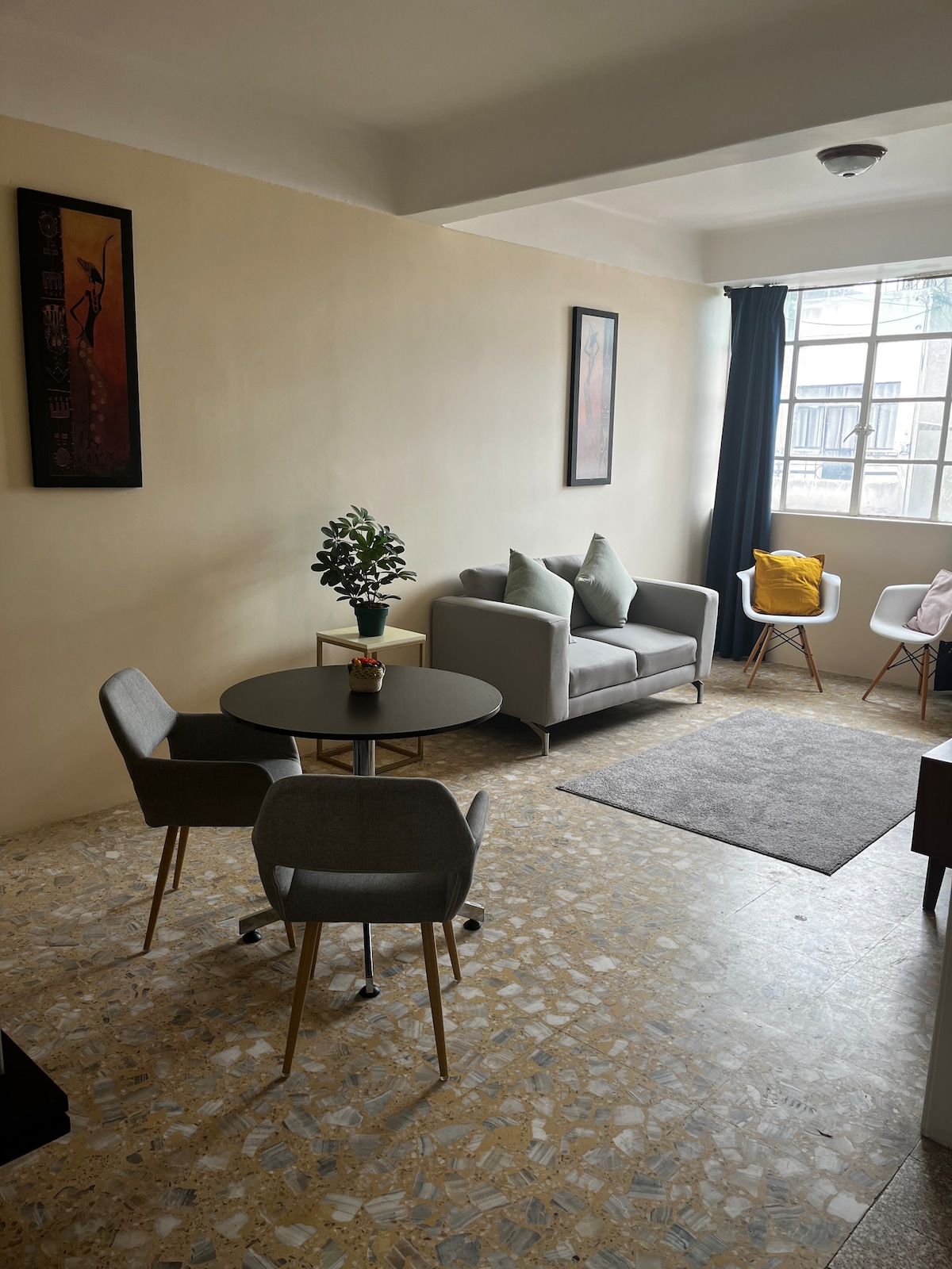 A modern living area includes a light-toned sofa adorned with decorative cushions, adjacent to a small round dining table with two gray chairs. A potted plant adds a touch of greenery. Natural light is welcomed through a large window with dark curtains.
