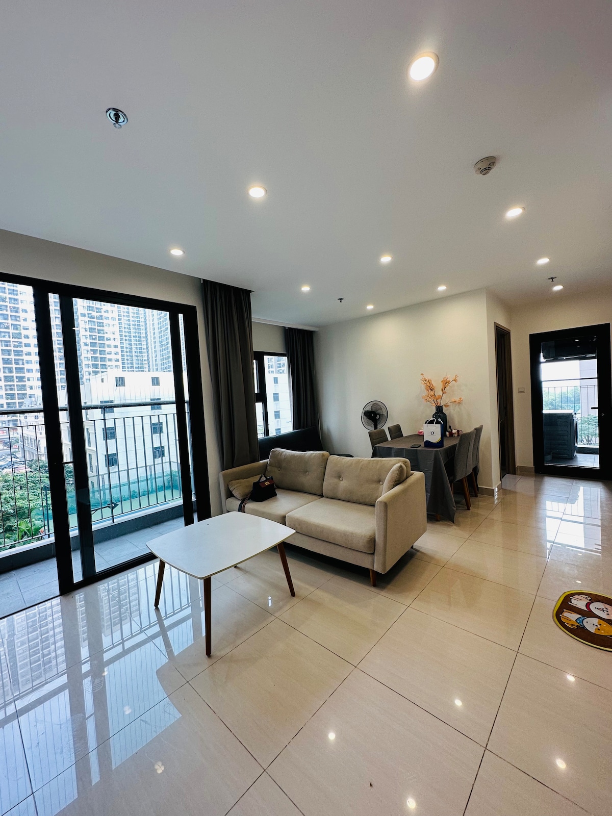 A modern living area features a comfortable sofa and a small coffee table. Large glass doors provide access to a balcony, allowing natural light to fill the space. Neutral-colored walls and tiled flooring contribute to a clean and open atmosphere.