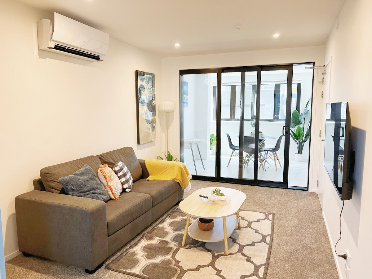 A modern living area features a comfortable grey sofa adorned with colorful cushions. A soft area rug lies beneath a round coffee table, which holds a small plant. Large glass doors open to an enclosed deck, allowing natural light to brighten the space.