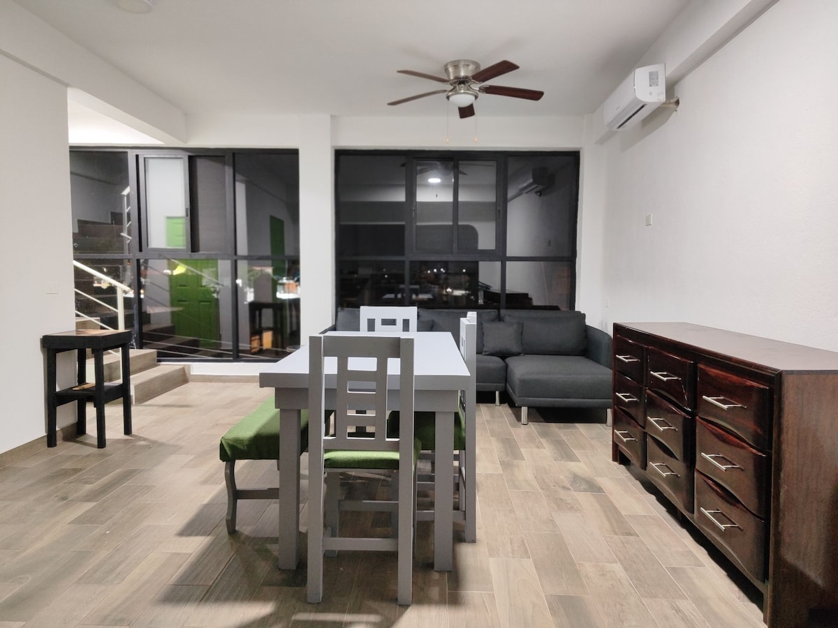 An open living area is presented, featuring a rectangular dining table with four chairs, a dark wooden dresser, and a small side table. A cozy sofa complements the space. Large windows allow natural light, while a ceiling fan is installed for comfort.