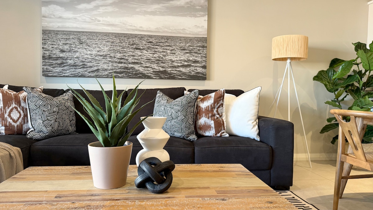 A comfortable living area features a dark sofa adorned with patterned cushions, complemented by a wooden coffee table displaying decorative items. A potted plant stands beside a white floor lamp, with a large black-and-white seascape photograph hanging above, enhancing the serene ambiance.