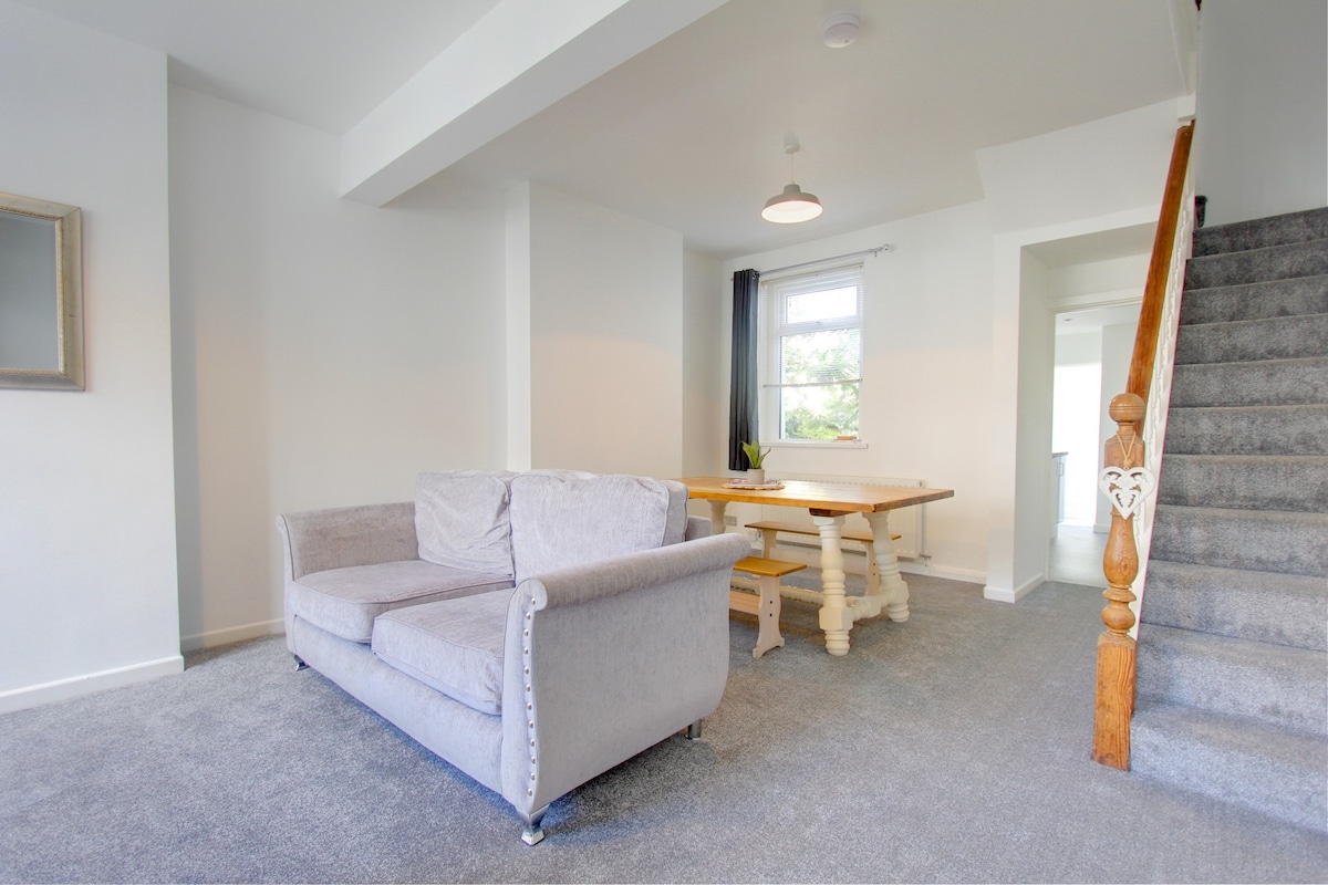 A spacious living area features a comfortable grey sofa and a large wooden dining table. Natural light enters through a nearby window, complementing the neutral decor. The staircase leads to the upper floor, while soft carpeting adds warmth to the space.