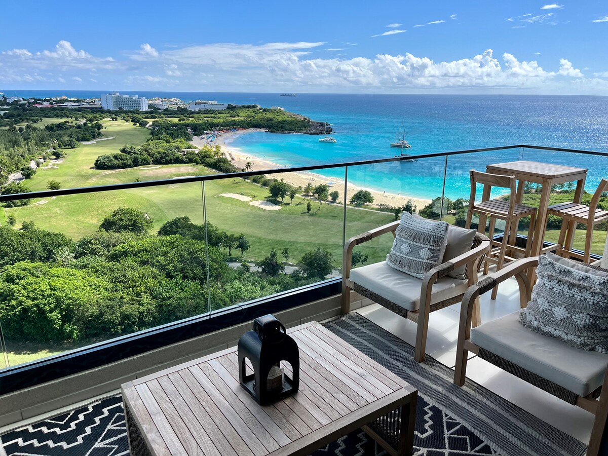 An inviting outdoor balcony showcases a stunning view of the ocean and surrounding landscapes. Comfortable seating, including two armchairs and a table, is positioned to enjoy the scenery. The space is adorned with a stylish patterned rug and a decorative lantern, creating a serene ambiance.