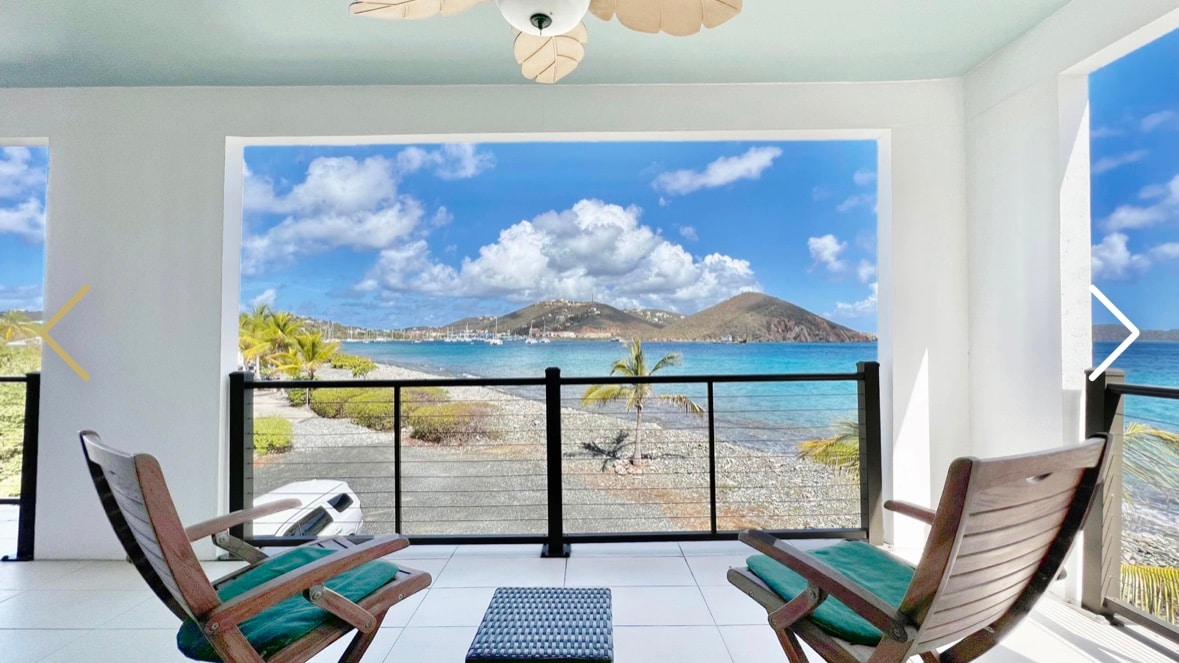 A spacious balcony offers panoramic ocean views and features two wooden chairs. A small table sits between them, while the vibrant blue water and distant hills create a serene backdrop. The ceiling is adorned with a decorative fan, enhancing the inviting atmosphere.