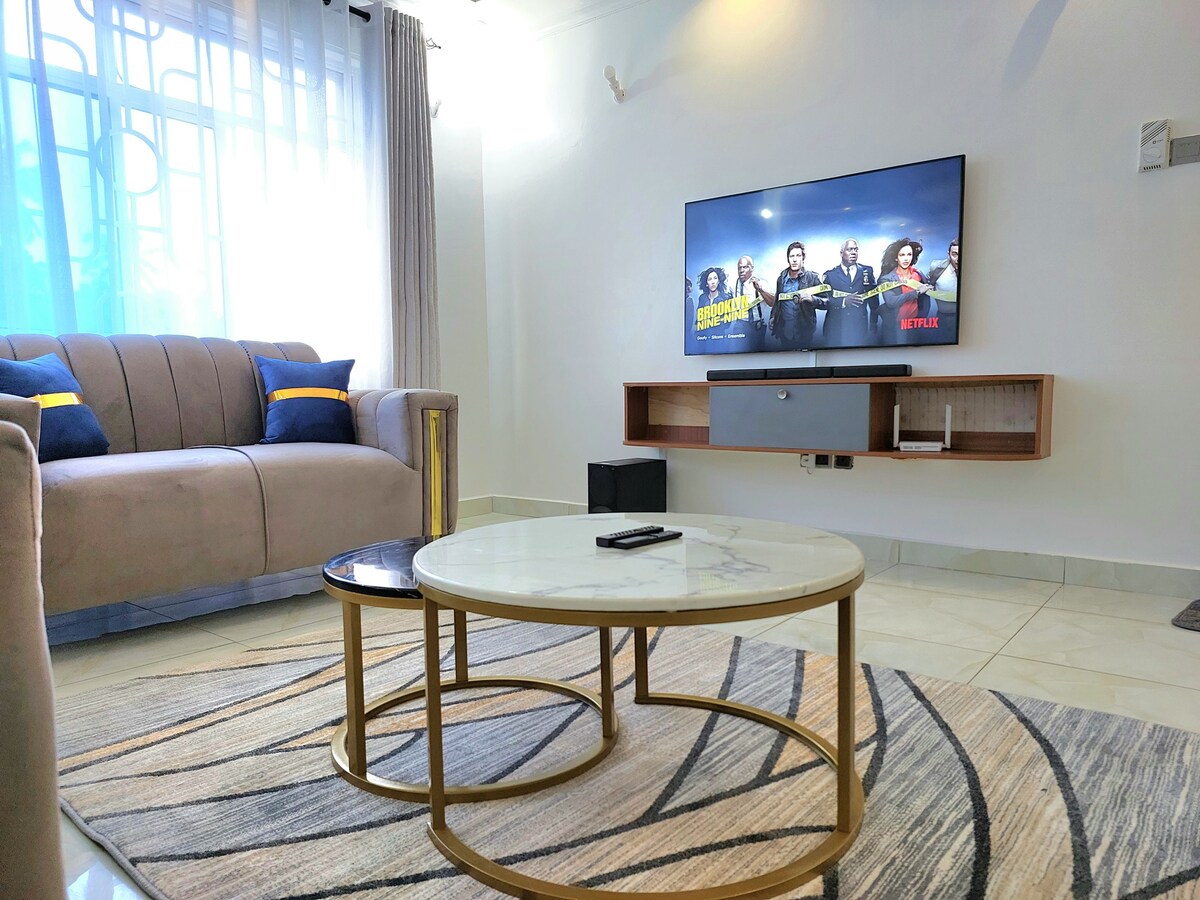 A modern living area features a plush sofa in neutral tones, accented by vibrant blue pillows. A marble coffee table with a circular design sits in front of the comfortable seating. A large flat-screen television is mounted on the wall, displaying content, while natural light filters through sheer curtains.