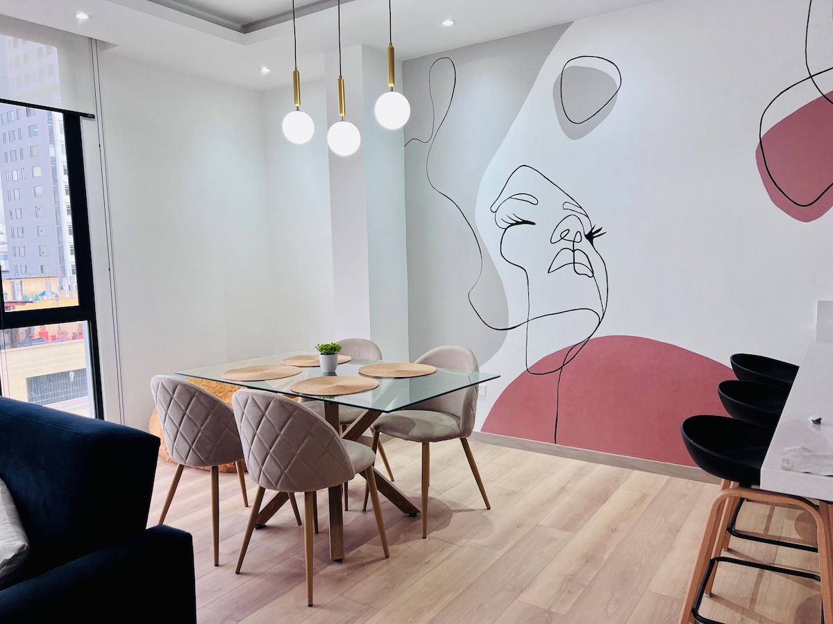 A modern dining area is featured, showcasing a glass table surrounded by six upholstered chairs. A line art mural adds a contemporary artistic touch to the wall. Minimalist pendant lights hang above, enhancing the bright and airy ambiance, complemented by natural light from the large window.