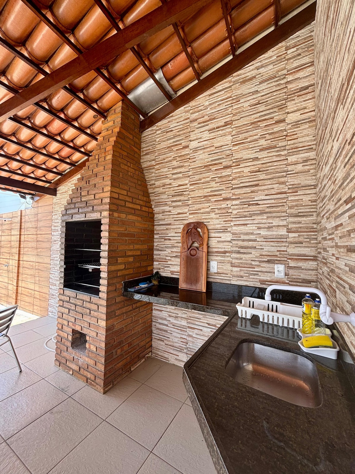 An outdoor area features a brick BBQ grill alongside a granite countertop containing a sink and essential cooking tools. Natural light illuminates the space, highlighting textured stone walls and a terracotta-tiled roof, creating a practical and enjoyable setting for al fresco dining.