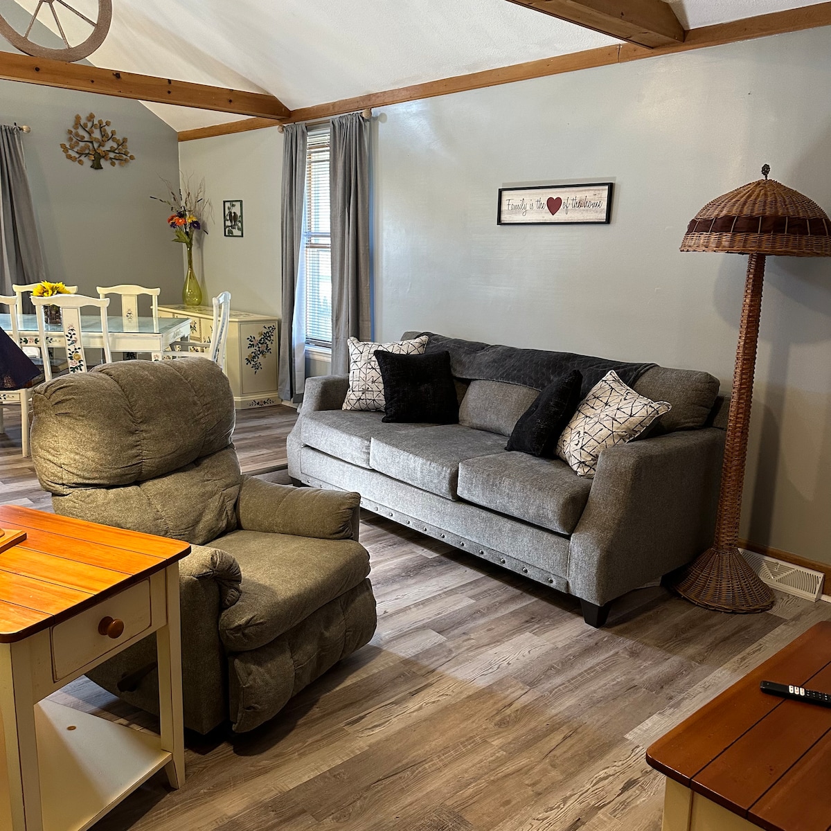 A comfortable living space features a gray sofa adorned with decorative pillows, complemented by a matching armchair. A wooden side table is positioned nearby, along with a tall lamp. Natural light enters through a window, illuminating the inviting atmosphere.