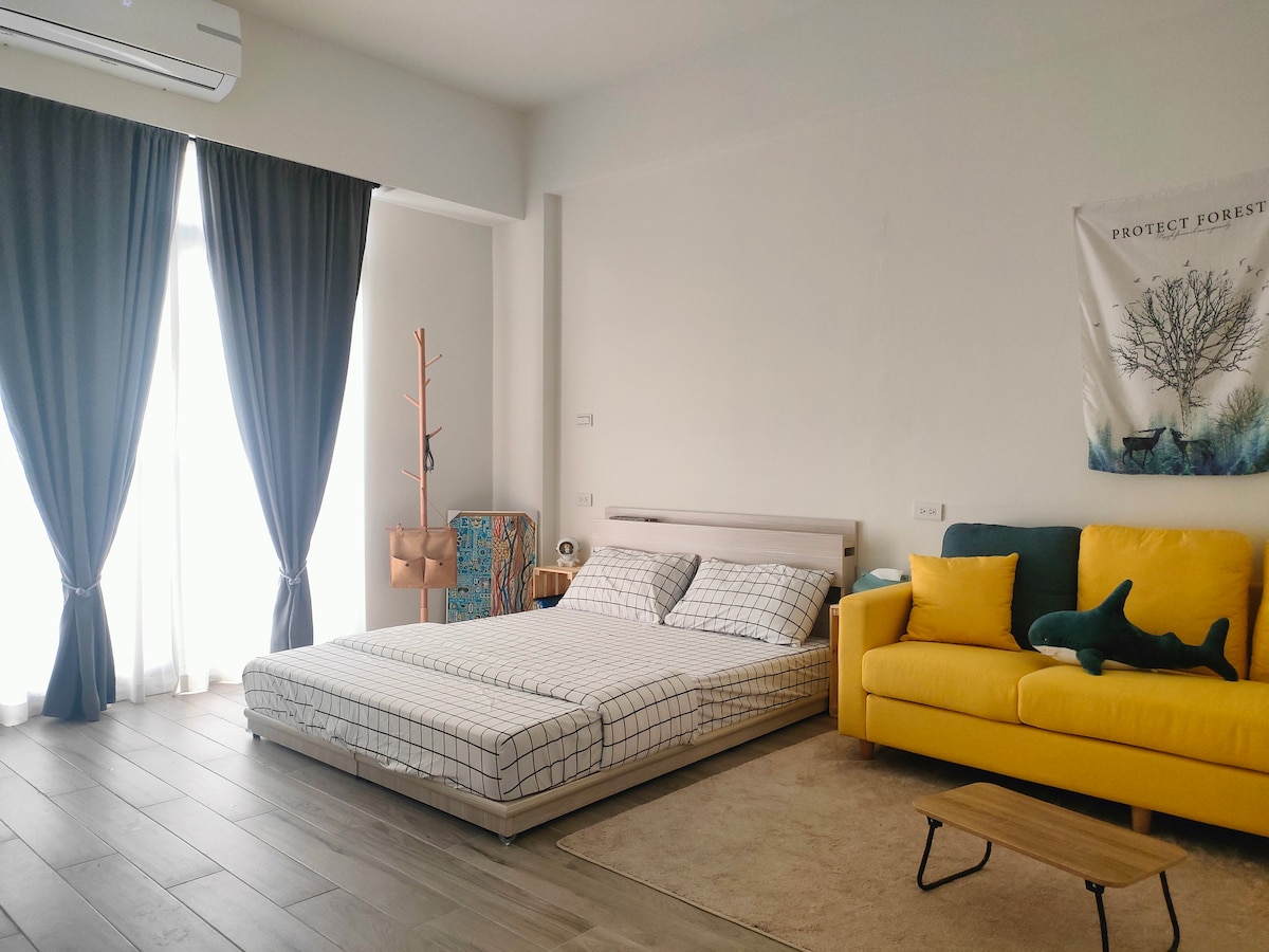 A spacious sleeping area features a low-profile bed adorned with checked bedding. Large, light-filtering curtains frame a window, providing ample natural light. A bright yellow sofa and a small wooden table complement the neutral tones of the room. A decorative wall hanging is visible in the background.