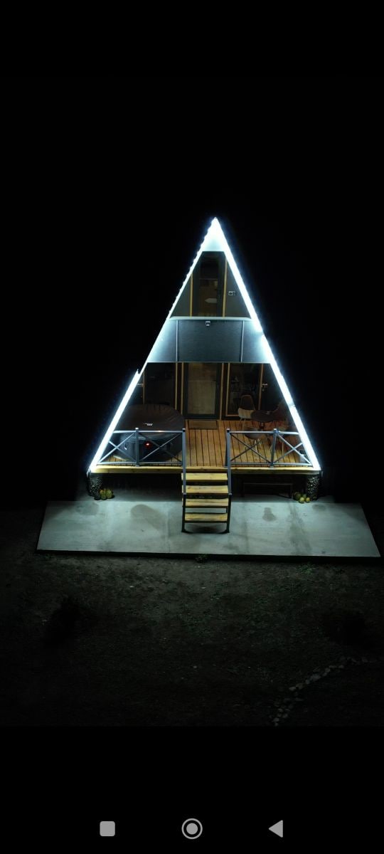 The triangular cabin is illuminated by bright LED lights, creating a striking contrast against the night sky. A welcoming entrance is framed by a wooden deck and is accessible via a set of steps. Surrounding greenery is visible on the ground.