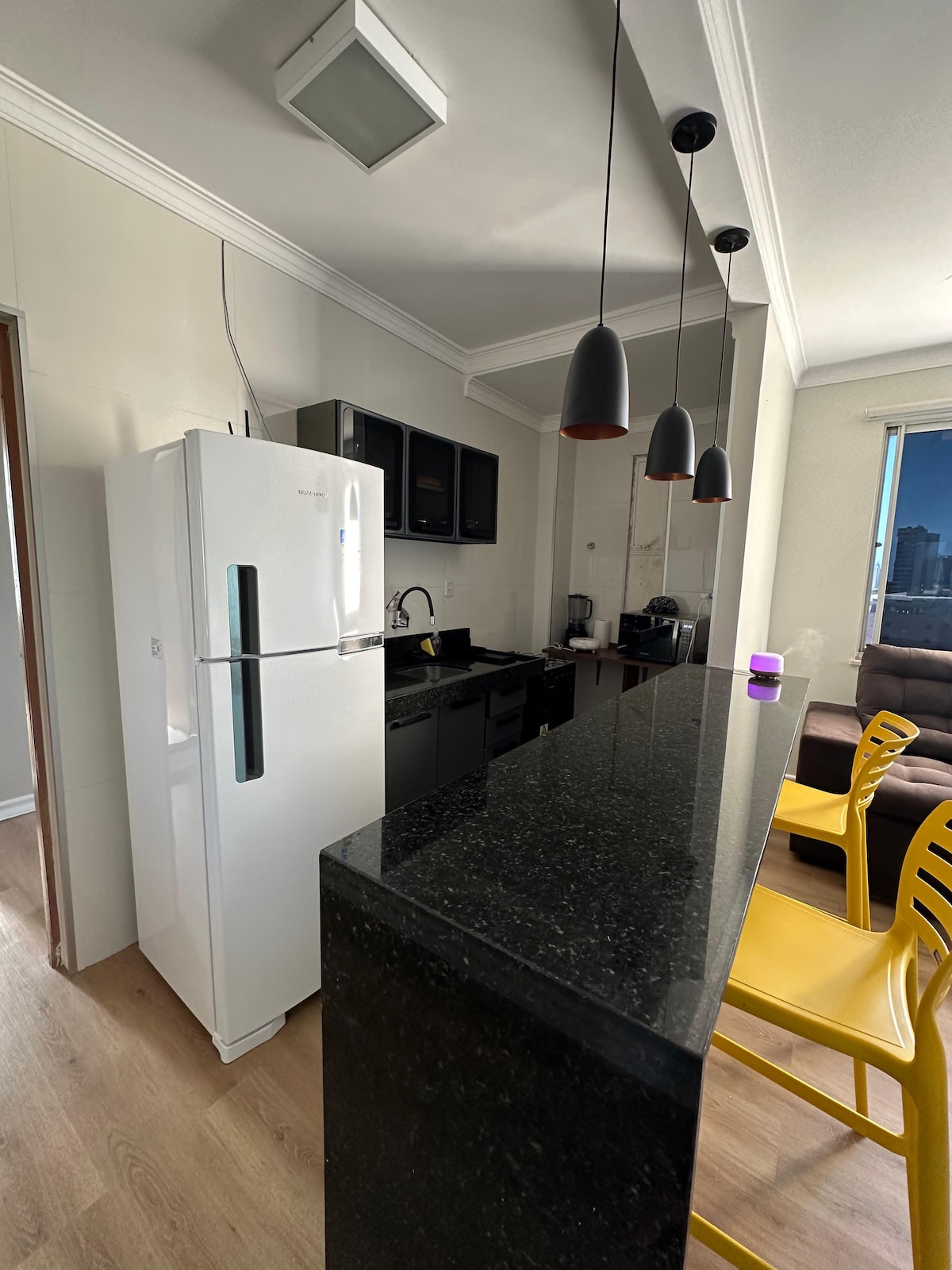 A modern kitchen features black cabinetry and a sleek granite countertop. Yellow bar stools are positioned at the counter, and a white refrigerator stands beside dark appliances. The bright space is illuminated by hanging light fixtures, and a window allows natural light to fill the room.