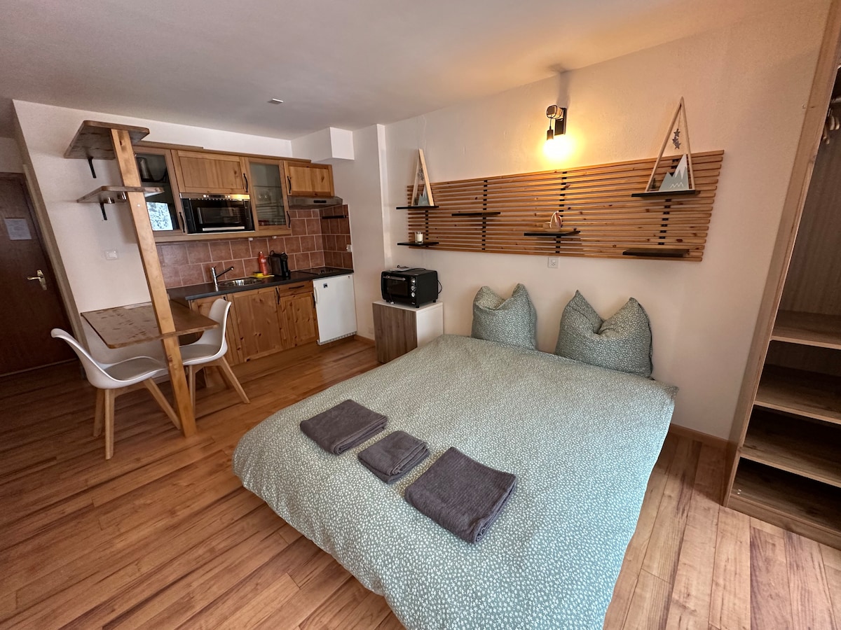 A spacious studio features a large bed with patterned bedding, complemented by neatly placed towels. An open kitchen area is visible, paired with a dining table and modern chairs. Wooden accents create a warm atmosphere, and natural light illuminates the space.