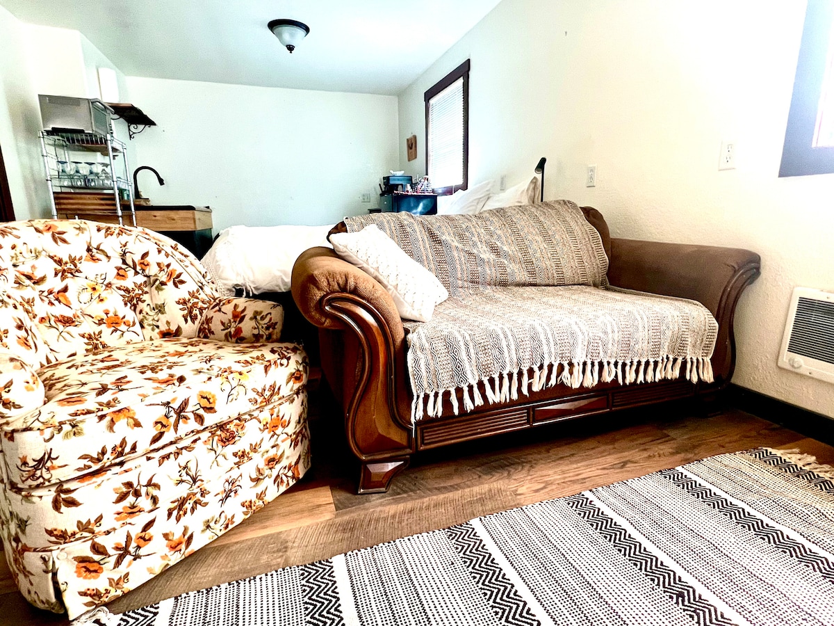 A cozy seating area features a floral-patterned armchair and a brown couch draped with a light blanket. A textured area rug lies below, complementing the wooden floor. Natural light enters through a nearby window, enhancing the inviting atmosphere.