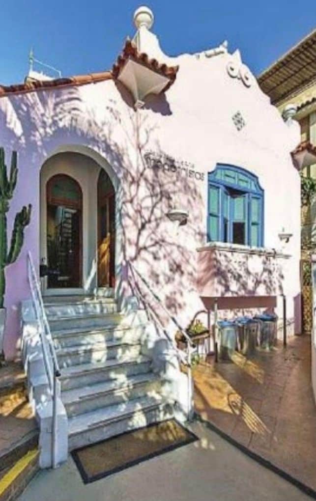 A charming colonial-style building features a pink exterior with ornate details and a staircase leading to the entrance. A blue window frame adds a pop of color, while shadows from nearby plants create an interesting texture on the facade.
