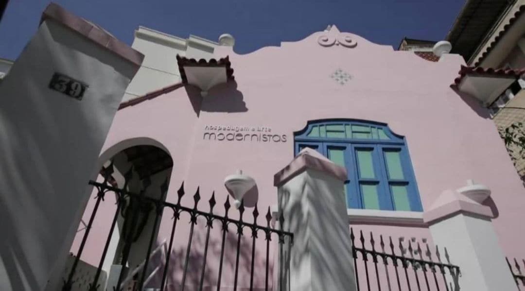 A colonial-style exterior is showcased with soft pink walls and intricate architectural details. Large blue windows add a vibrant contrast, while a gated entrance provides charm. The building's name, 'modernistas', is prominently displayed, inviting guests to enjoy the artistic atmosphere.