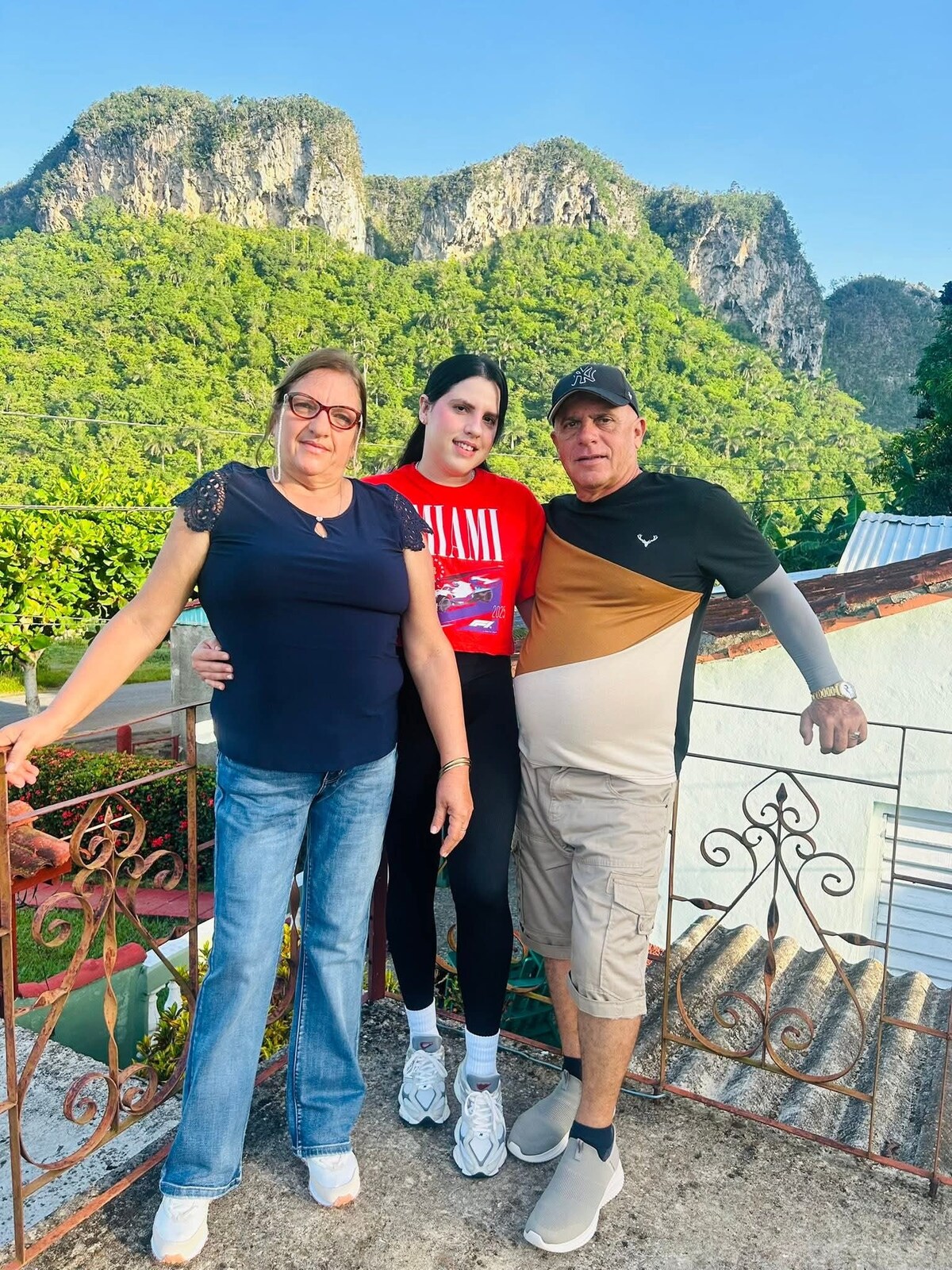 A group of three individuals stands together on a terrace with a scenic backdrop of lush green hills and rocky cliffs. The figures are casually dressed, enjoying the natural beauty surrounding them. The terrace railing features ornamental wrought iron details.
