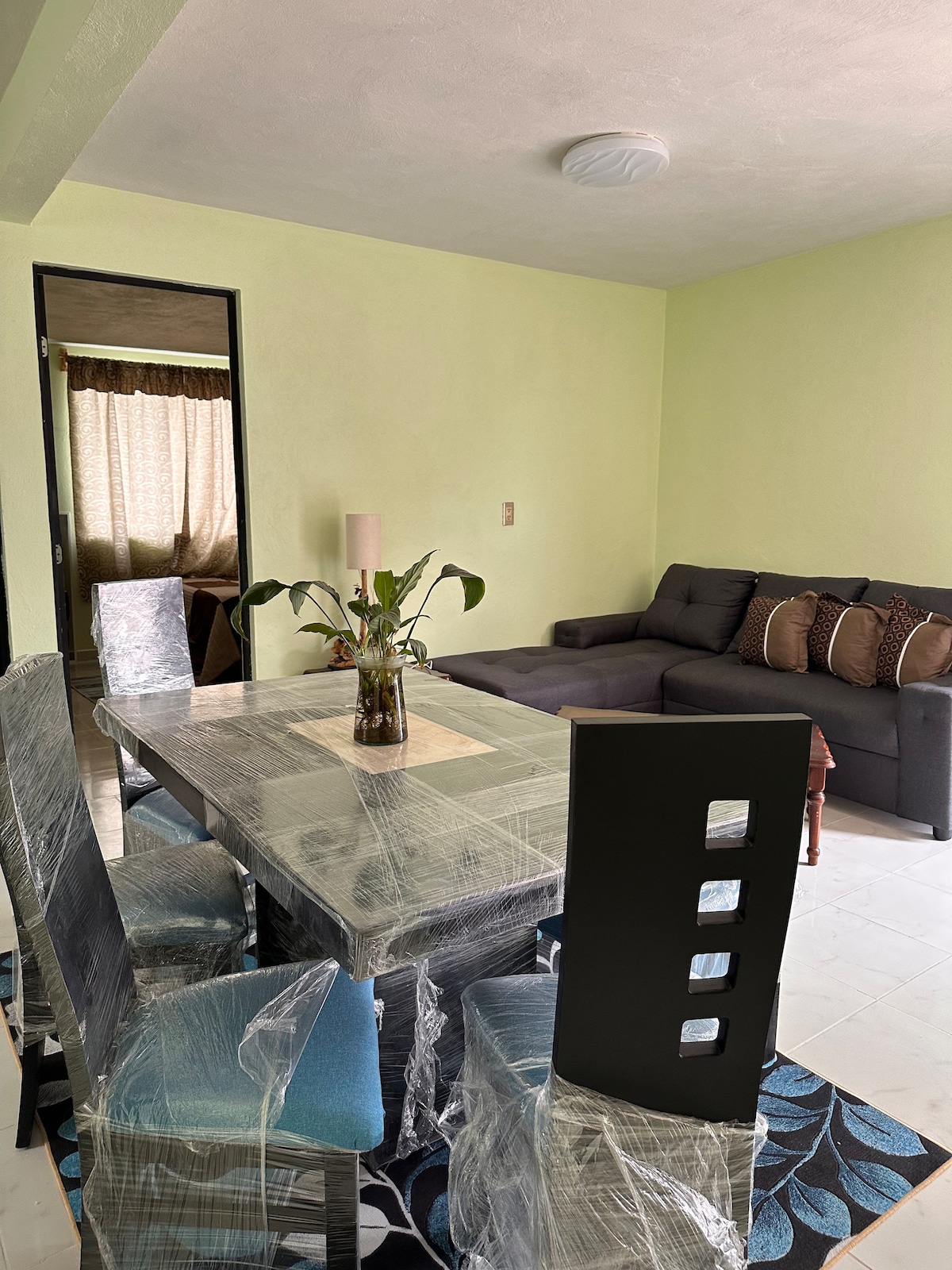 A dining area is presented with a table surrounded by four chairs, wrapped in protective material. A floral centerpiece is placed on the table, with a cozy seating area visible in the background, featuring a dark gray sofa paired with decorative pillows.