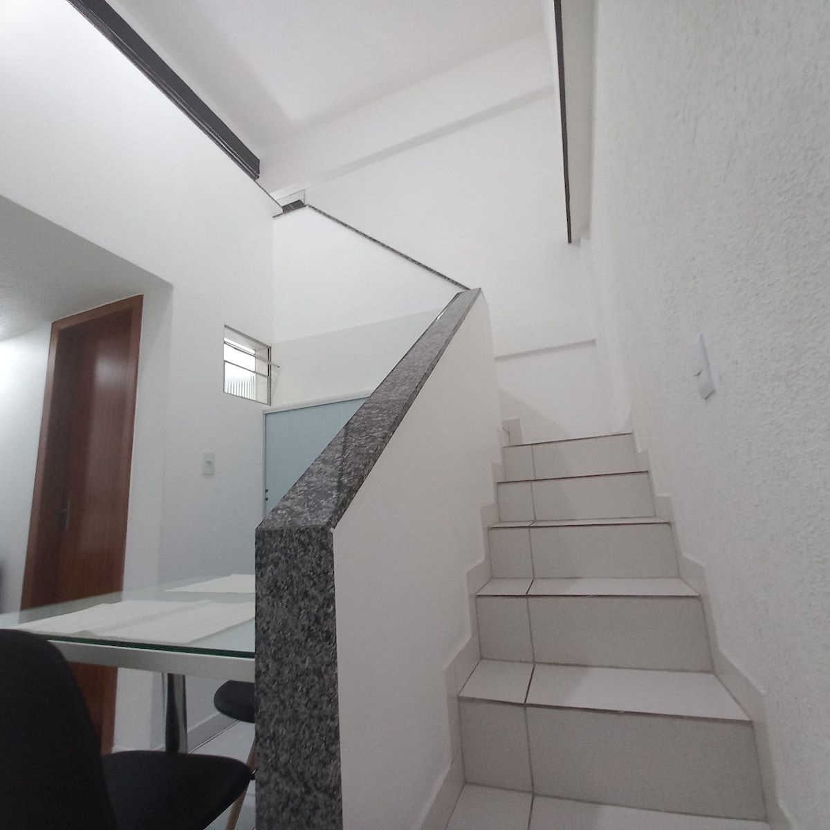 A well-lit stairway with white tiled steps leads to an upper level. The walls are painted white, creating a bright atmosphere. A glass-top table is visible in the foreground, complemented by a black chair.
