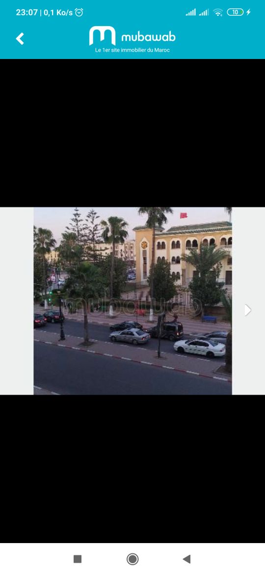 A view of a street lined with palm trees and parked cars. A building with distinctive architecture and large windows is visible in the background, accompanied by a flag. The scene captures an early evening atmosphere with soft lighting.